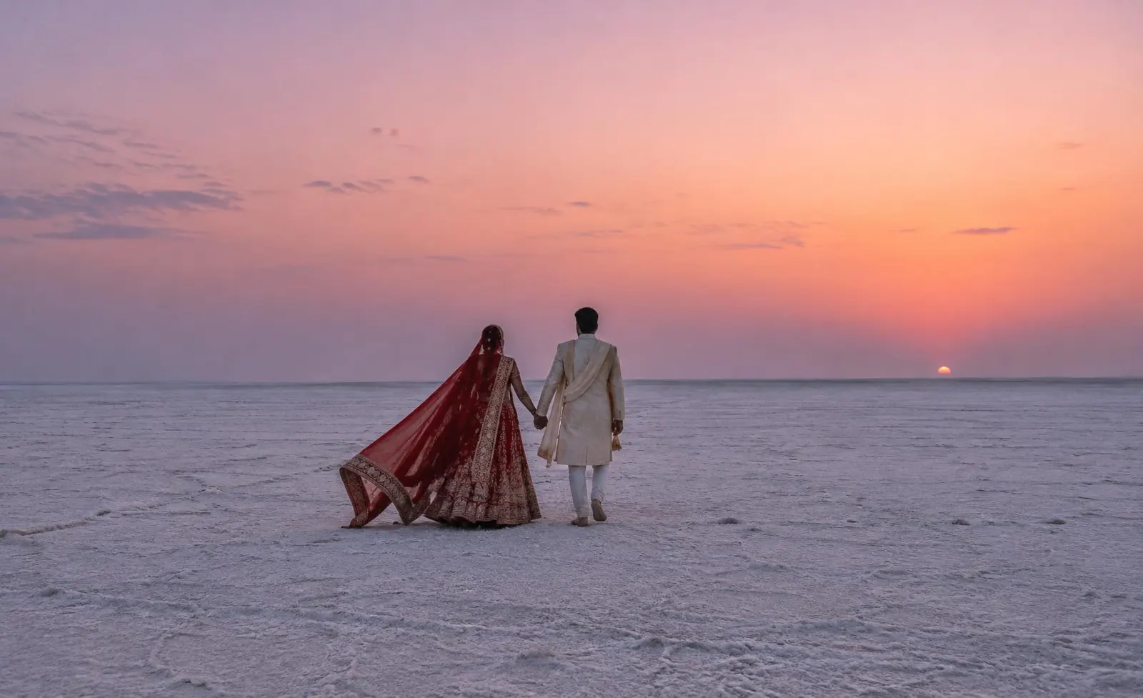 A bride and groom walking hand in hand across a white salt desert at sunset